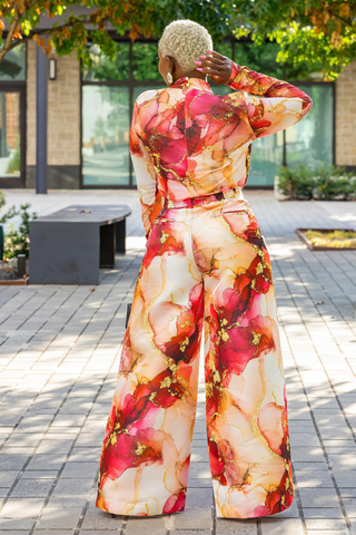 A woman stands outdoors on a paved walkway, viewed from behind, wearing a long-sleeve wide-leg jumpsuit with a bold abstract floral print in red, pink, orange, and cream tones.