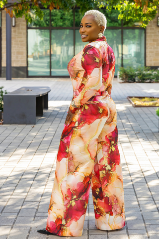 A woman with short blonde hair is shown from the back wearing a high-neck, wide-leg jumpsuit with an abstract floral print in red, pink, beige, and gold. She is standing on a paved outdoor walkway.