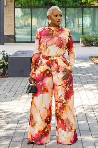 A woman stands outdoors on a paved walkway wearing a matching long-sleeve high-neck jumpsuit with a bold abstract floral print in pink, red, peach, and gold tones. She is holding a small black handbag.