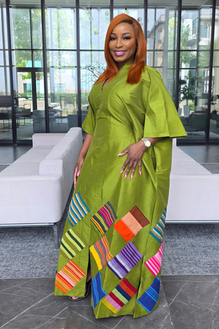A woman with orange hair wearing a bright olive green traditional dress with colorful geometric patterned panels at the hem, standing in a modern lobby with glass windows and white sofas.