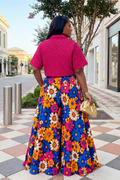 Woman wearing a bright pink top and colorful floral skirt in an urban setting.