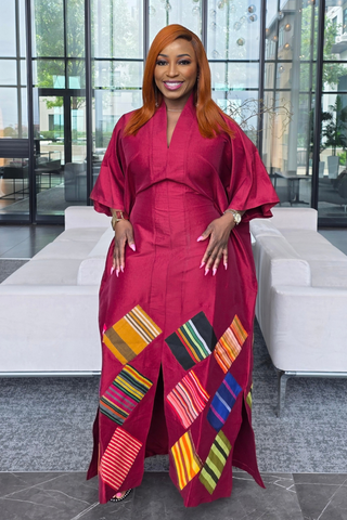 A woman with red hair wearing a vibrant magenta African dashiki dress with colorful geometric patterned panels, standing in a modern lobby with white sofas and glass windows.
