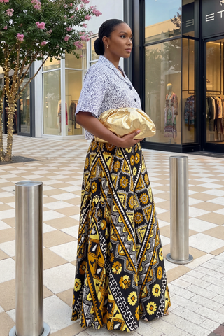 Woman in a patterned skirt and blouse holding a gold clutch in front of a store.