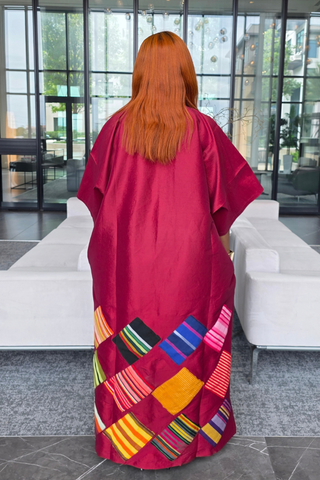 A person viewed from behind wearing a long magenta kaftan dress with a colorful striped woven patchwork design along the lower hem, standing in a modern indoor lobby with white sofas and large glass windows.