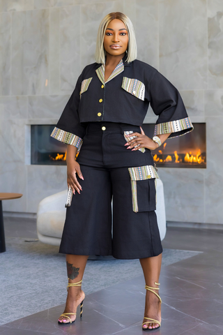 A woman models a black two-piece matching set consisting of a cropped button-up top and wide-leg cropped pants, both featuring beige and multicolor striped trim details. She is standing in a modern indoor setting with a fireplace in the background.