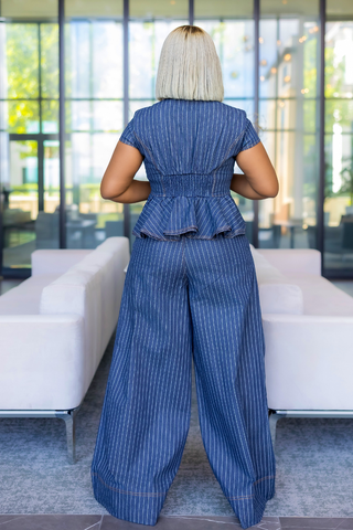 A woman viewed from behind wearing a blue pinstriped short-sleeve jumpsuit with a smocked waist and wide legs, standing in a modern room with white sofas and large windows.