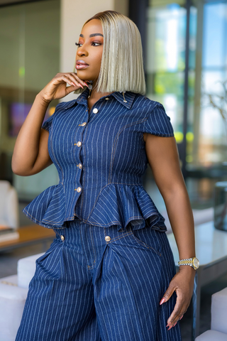 A woman wearing a blue pinstriped matching set consisting of a short-sleeve button-front peplum top and wide-leg pants, posed in a modern indoor setting with blurred windows in the background.