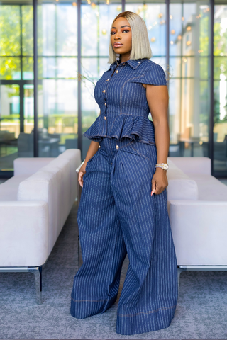 A woman with platinum blonde hair models a blue and white pinstriped short-sleeve button-up shirt and matching wide-leg pants set in a modern office lobby with white sofas.