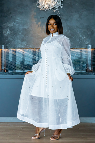 Woman wearing a white dress standing indoors with a textured wall and chandelier in the background.