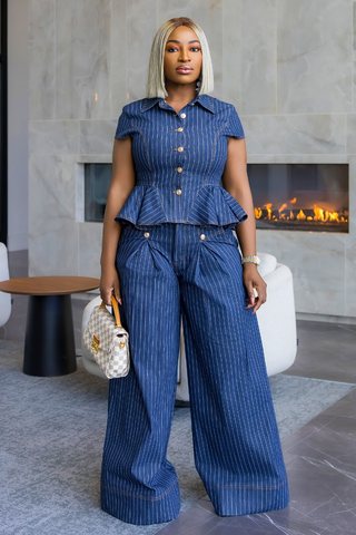 A woman stands in a modern indoor setting wearing a matching blue pinstriped short-sleeve button-front top and wide-leg pants set, holding a small patterned handbag.