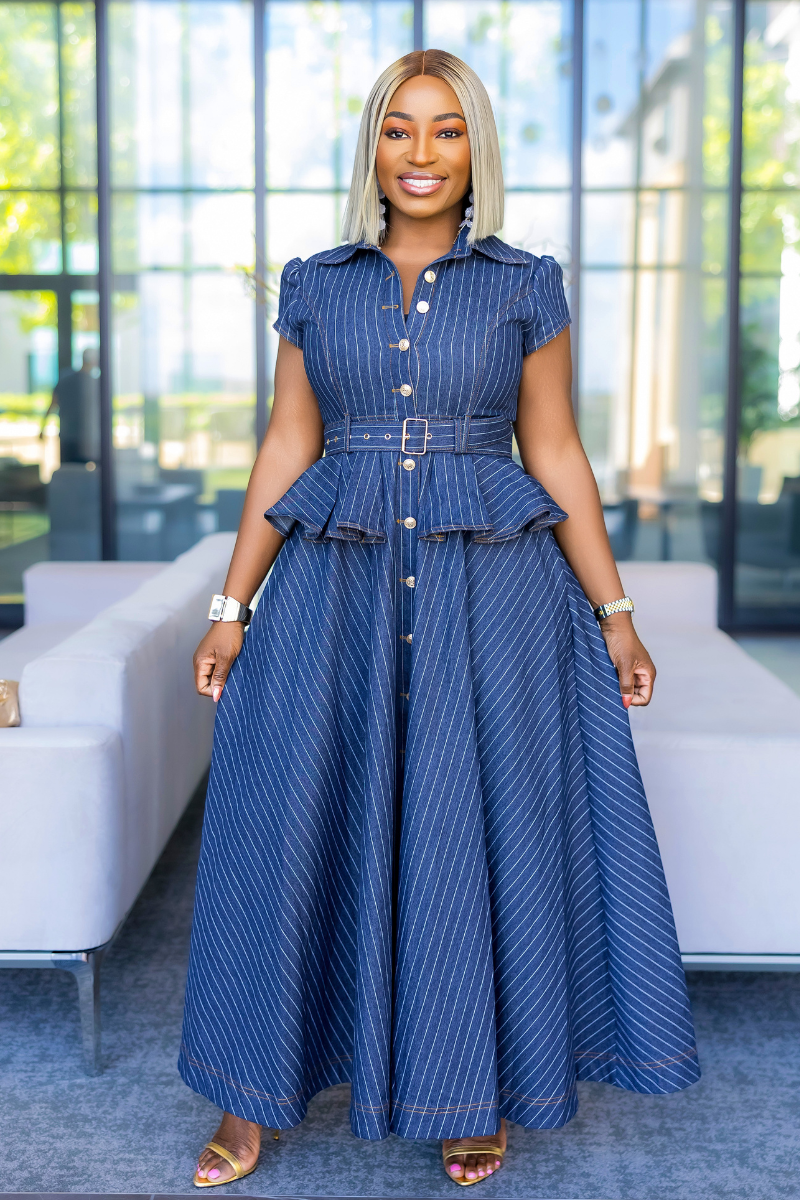 A woman modeling a blue pinstriped maxi dress with a button-up front, short sleeves, and a matching belt with a silver buckle, standing in a modern indoor setting with a white sofa and large windows in the background.