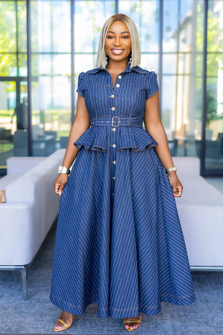 A woman modeling a blue pinstriped maxi dress with a button-up front, short sleeves, and a matching belt with a silver buckle, standing in a modern indoor setting with a white sofa and large windows in the background.