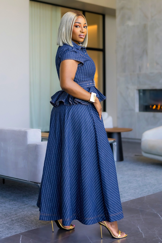 A woman wearing a blue pinstriped maxi dress with a ruffled high-neck bodice and matching belt, styled with gold high heels in a modern living room setting.