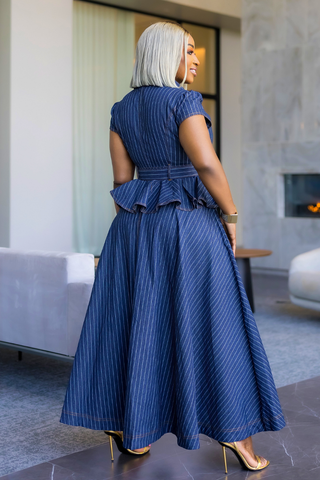 A woman with platinum blonde hair is shown from behind wearing a navy blue pinstriped maxi dress with a peplum waist, styled with clear high-heeled sandals in a modern living room setting.