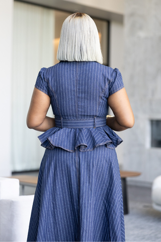 A woman with a platinum blonde bob is shown from behind wearing a blue pinstriped dress with puff sleeves and a ruffled peplum waist, set in a modern indoor environment.