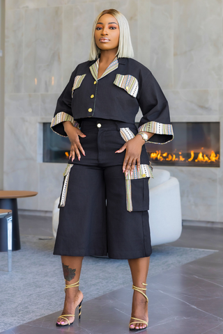A woman modeling a black two-piece outfit consisting of a cropped button-front top with striped trim and matching high-waisted cargo-style shorts, photographed in a modern indoor setting with a fireplace in the background.
