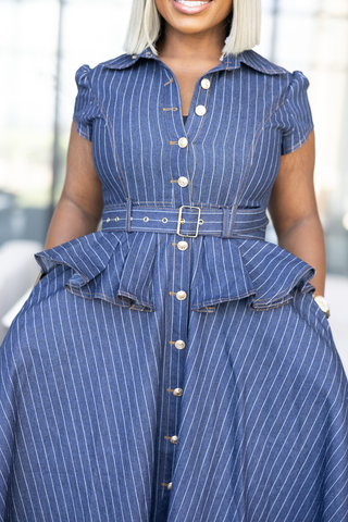 A close-up of a woman wearing a blue pinstriped shirt dress with a matching belt and silver buckle, featuring short sleeves, a button-down front, and a peplum waist.