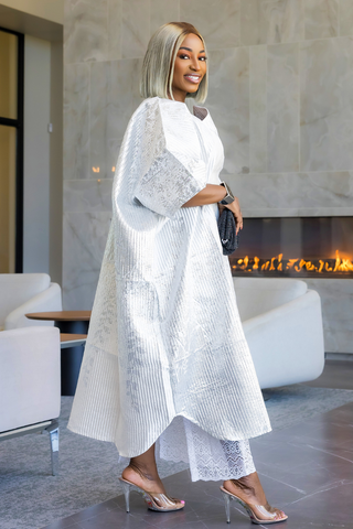 A woman models a white long-sleeve kaftan dress with intricate lace detailing, paired with matching white lace pants and clear high-heeled sandals, standing in a modern living room with a fireplace.