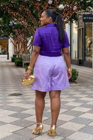 Woman in purple top and light purple shorts holding a gold clutch on a city street.