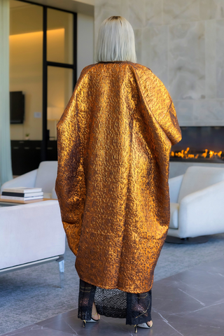 A person viewed from behind wearing a voluminous, textured metallic gold robe in a modern living room with a fireplace and white armchairs.