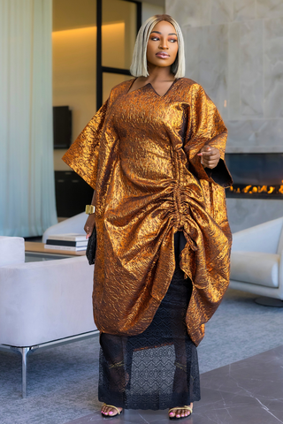 A woman with platinum blonde hair wearing a shimmering metallic bronze kaftan dress with a gathered front, standing in a modern living room setting.
