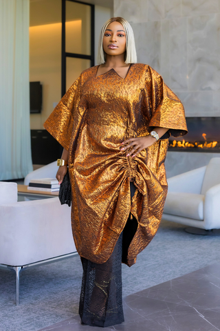 A woman with platinum blonde hair models a metallic gold kaftan dress with a gathered, draped design and wide sleeves, paired with black sheer wide-leg pants in a modern living room setting.