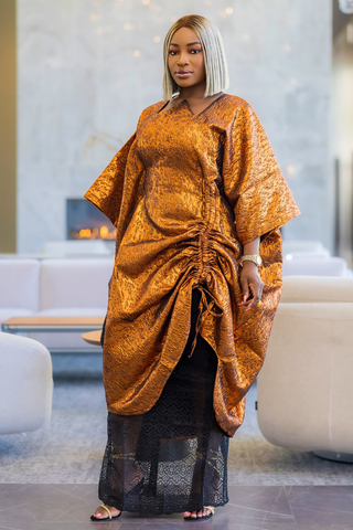A woman wearing a shimmering metallic gold kaftan dress with a black lace skirt, standing in a modern living room with white sofas and a fireplace.
