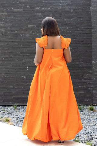 Woman wearing an orange dress with ruffled sleeves against a dark brick wall.
