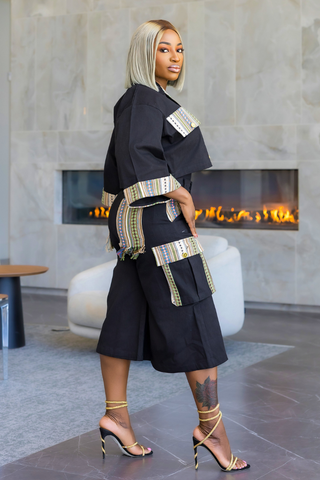 A woman with platinum blonde hair models a black African wax print (Ankara) dress with colorful patterned trim in a modern lobby with a fireplace in the background.