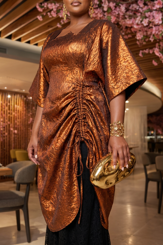 Woman in a bronze dress holding a gold clutch in an indoor setting with floral decorations.