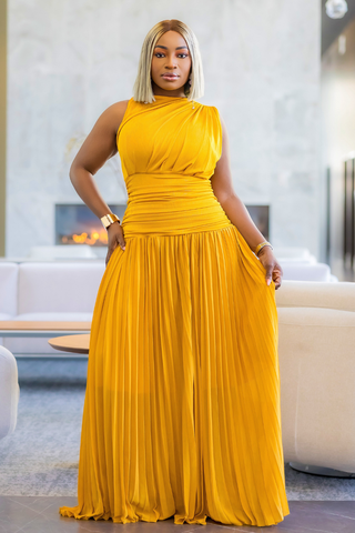 A woman wearing a sleeveless, high-neck, floor-length yellow dress with a pleated skirt, standing in a modern living room with a fireplace and white sofas in the background.