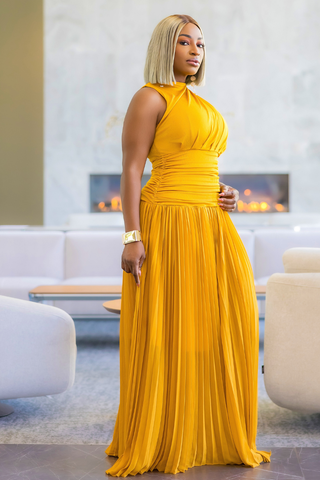A woman wearing a sleeveless, high-neck, floor-length yellow pleated gown, standing in a modern living room with a fireplace and white sofas in the background.