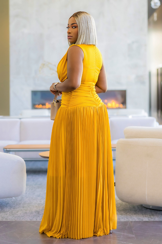 A woman wearing a sleeveless mustard yellow pleated maxi dress, standing in a modern living room with a fireplace and white sofas in the background.