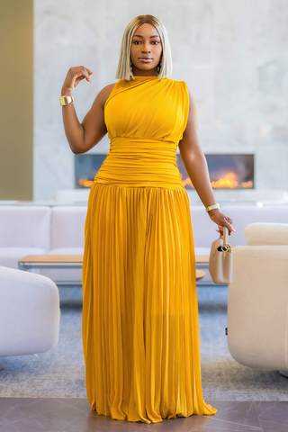 A woman modeling a sleeveless, one-shoulder yellow maxi dress with a ruched bodice and pleated skirt, standing in a modern living room setting.