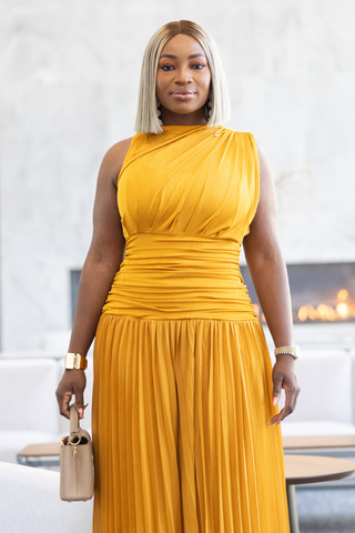 A woman wearing a mustard yellow one-shoulder pleated dress, holding a small beige handbag, standing in a modern living room setting.