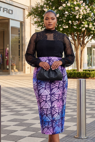 Woman in a black top and purple patterned skirt standing outdoors near a building.