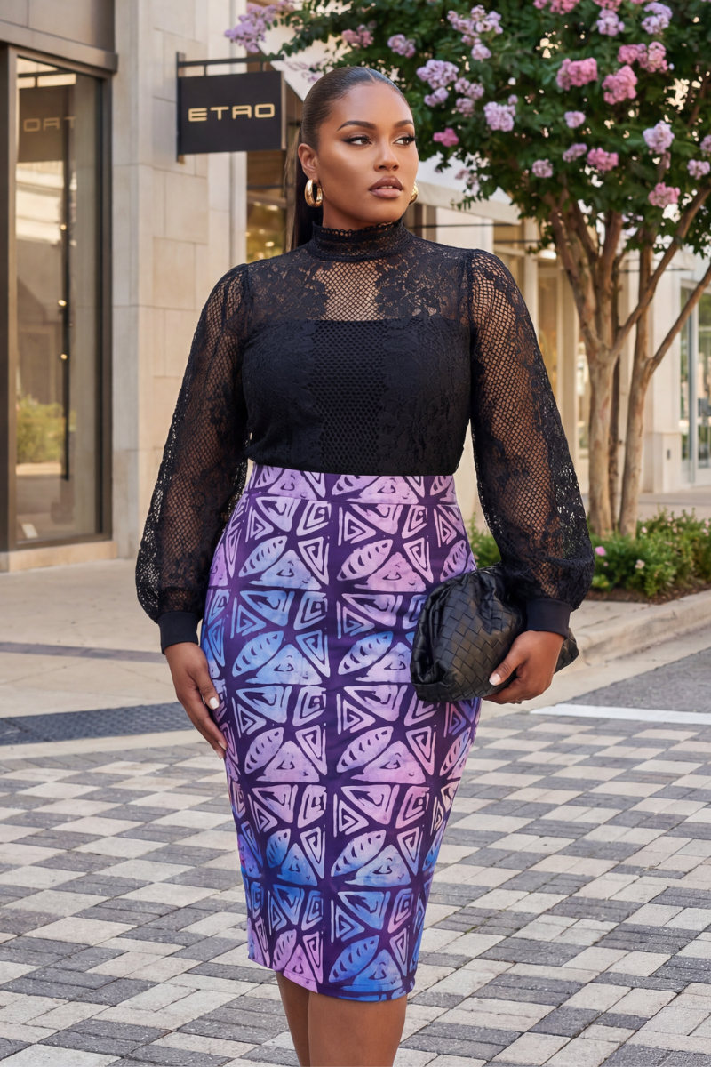 Woman wearing a black lace top and patterned skirt standing on a sidewalk with a building and tree in the background.