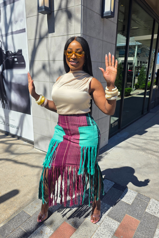 Woman in a colorful skirt and beige top waving in front of a building.