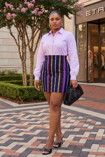 Woman in a pink shirt and striped skirt standing on a patterned sidewalk with a store entrance in the background.