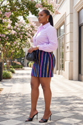 Woman in a striped skirt and light purple shirt standing on a sidewalk with trees and buildings in the background.