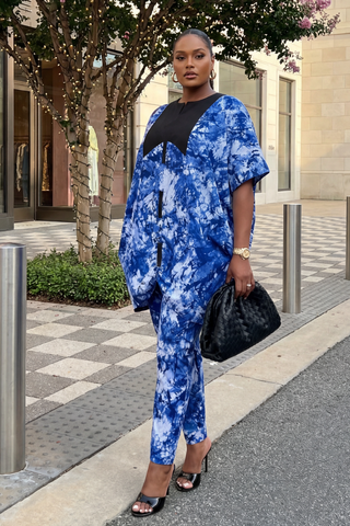 Woman in a blue tie-dye outfit standing on a sidewalk with trees and buildings in the background.