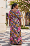 Woman wearing a colorful patterned dress standing outdoors near a building with pink flowers.