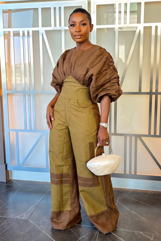 A woman models a two-piece brown and olive green outfit consisting of a voluminous short-sleeve top and matching wide-leg trousers, holding a small white handbag.
