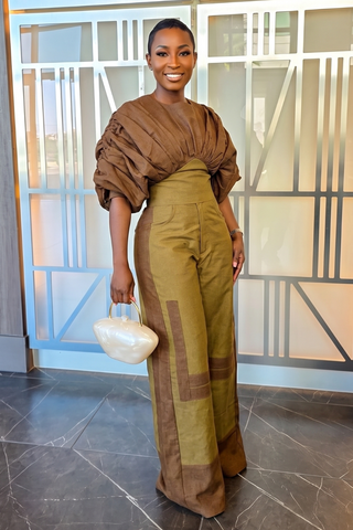 A woman stands indoors wearing a brown and olive green patchwork jumpsuit with dramatic draped sleeves and wide legs, holding a small white handbag. The background shows a modern glass-paneled wall and dark tiled floor.
