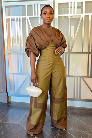 A woman stands indoors wearing a brown and olive green two-piece outfit with a ruched top and wide-leg pants, holding a small white handbag.
