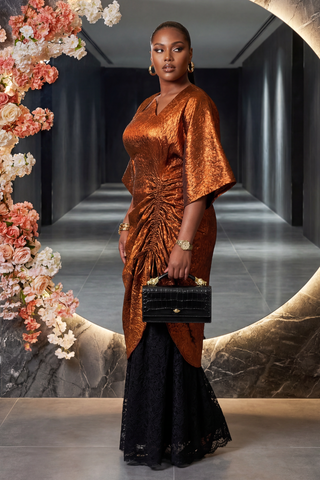 Woman in a bronze dress holding a black handbag against a decorative floral and stone wall.