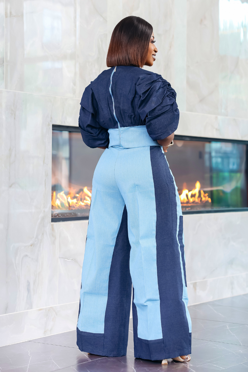 A woman modeling a light blue and navy color-blocked jumpsuit with dramatic puffed sleeves, standing in front of a modern fireplace.