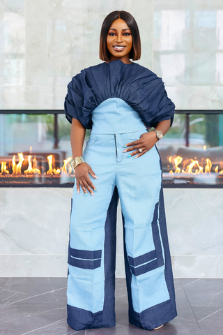 A woman modeling a light blue and navy color-blocked jumpsuit with dramatic puffed sleeves, standing in front of a modern fireplace.