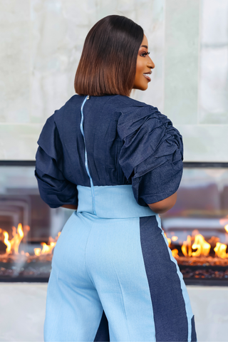 A woman shown from the back wearing a two-tone jumpsuit in navy and light blue, standing in front of a fireplace.