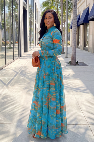 Woman in a blue floral dress standing on a sidewalk with palm trees and buildings in the background.